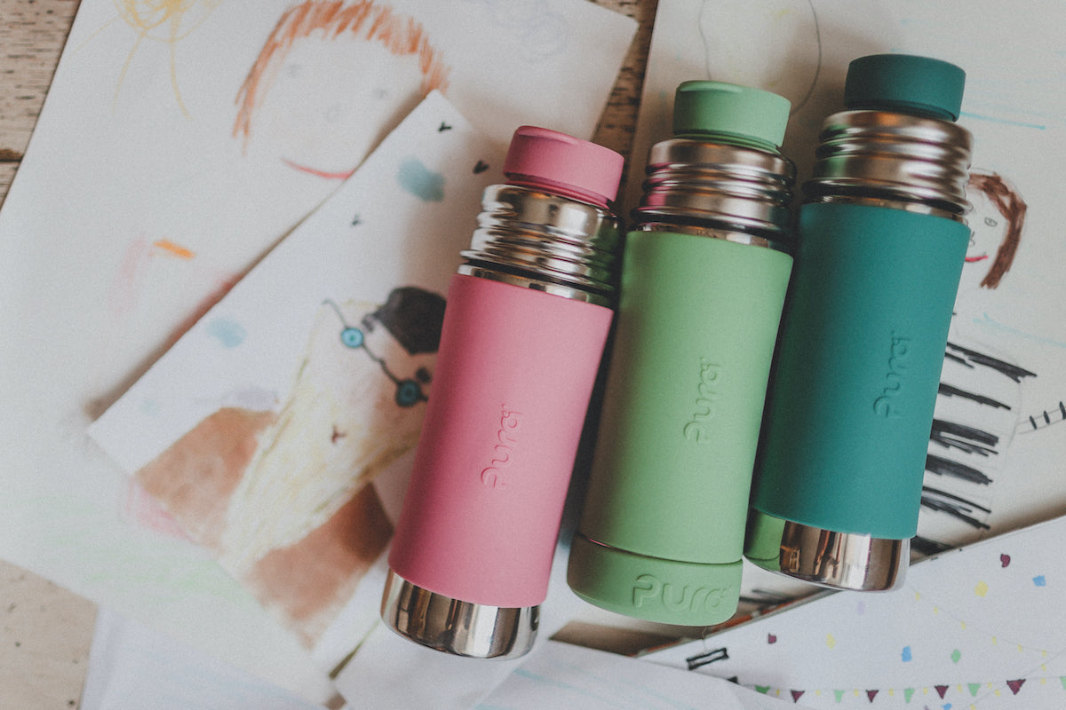 Three colorful Pura Stainless water bottles on top of a child’s drawings.