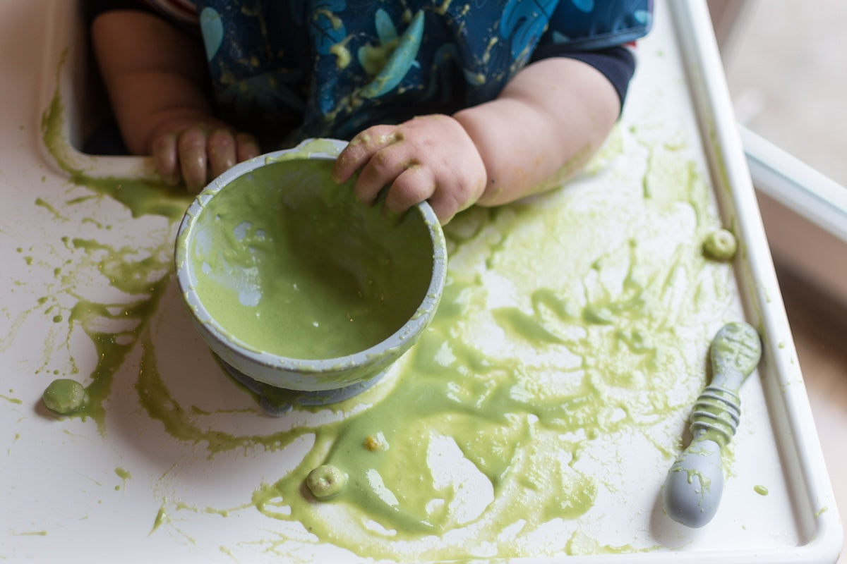 Baby eating with messy green food all over silicone bowl and spoon